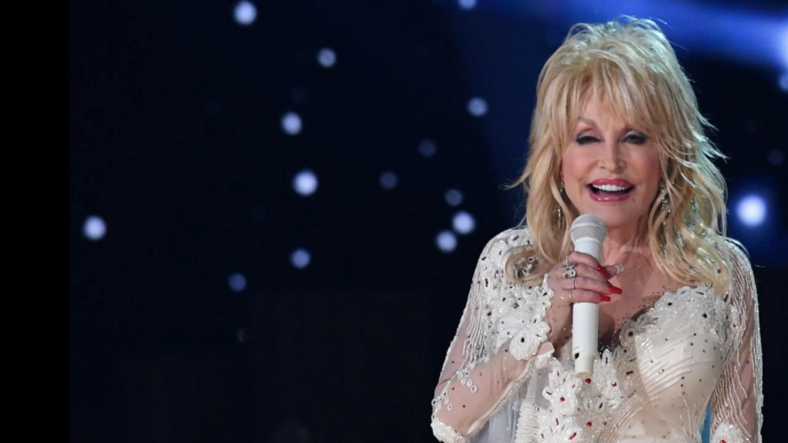 Dolly Parton onstage in an embellished white outfit, holding a microphone