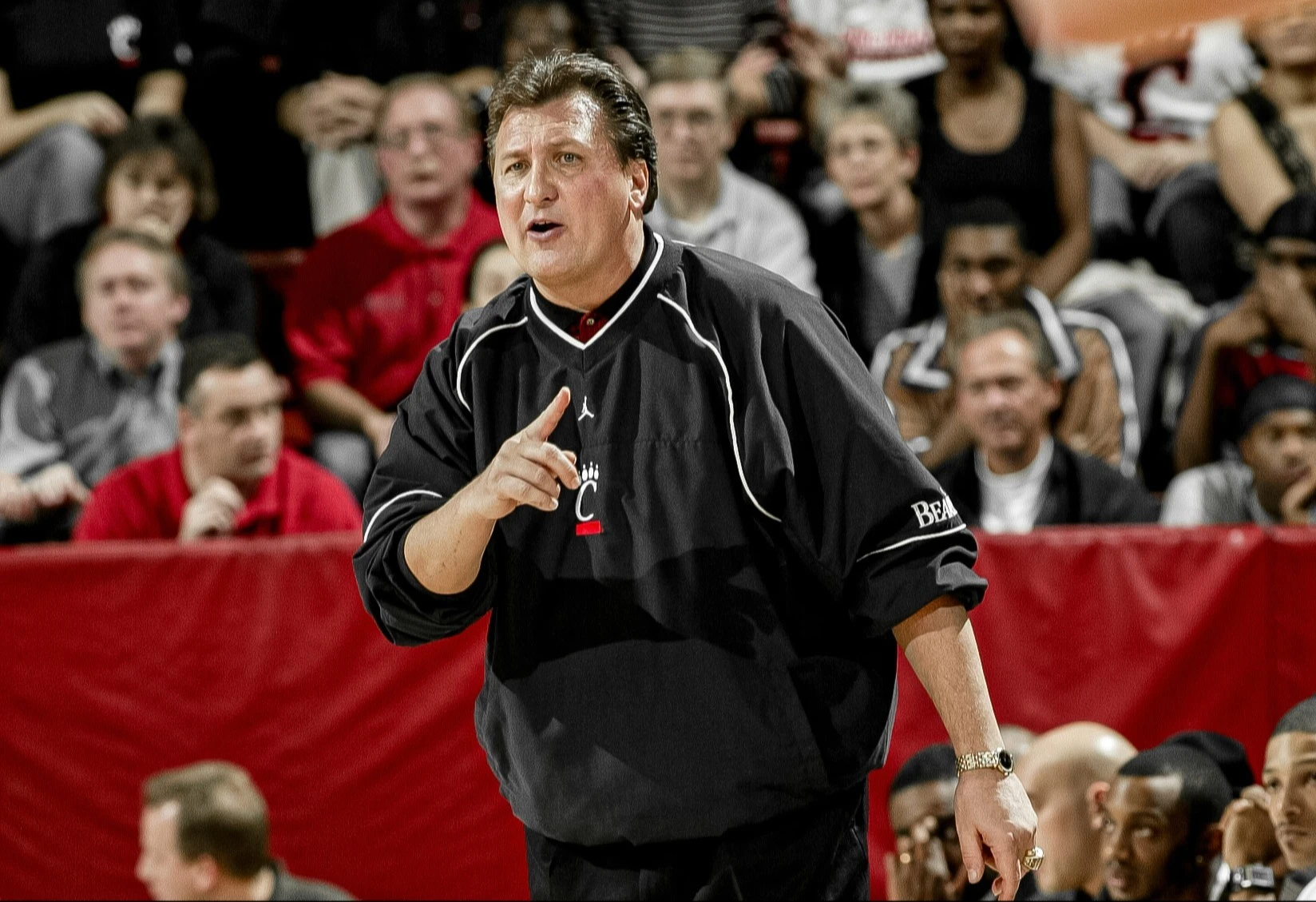 Bob Huggins in a black Cincinnati Bearcats pullover gesturing from the sideline during a game in front of fans