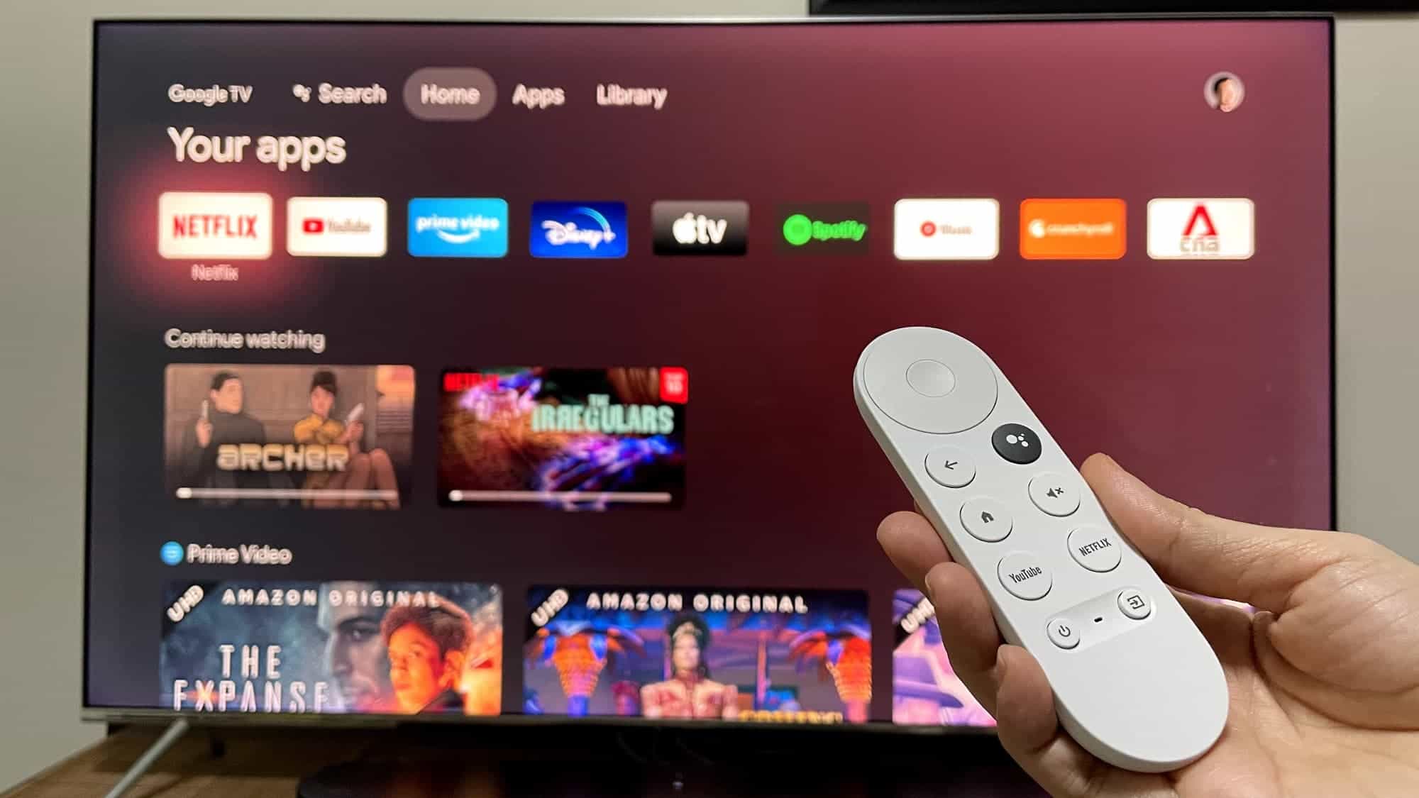 A Google TV voice remote held in front of a television displaying an Android-based Google TV home screen