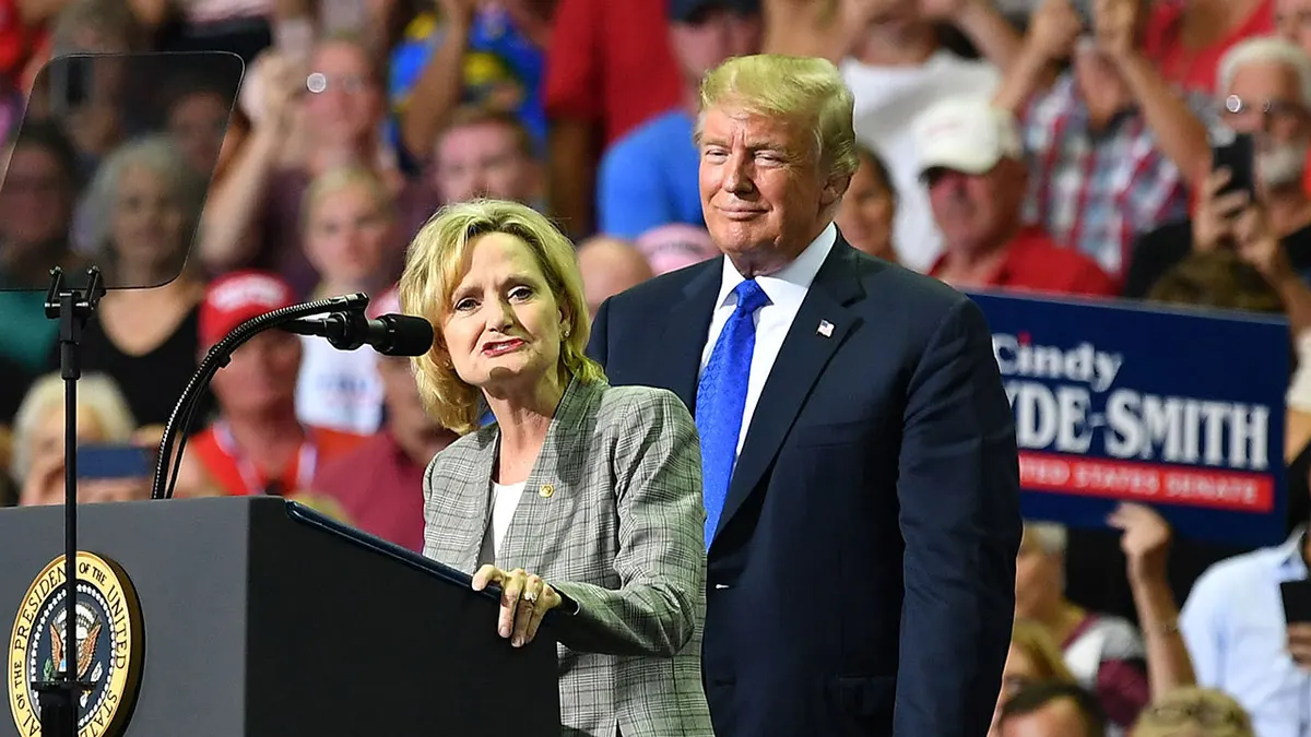 Cindy Hyde-Smith speaks at a campaign podium while Donald Trump stands behind her in an earlier Mississippi rally photograph.