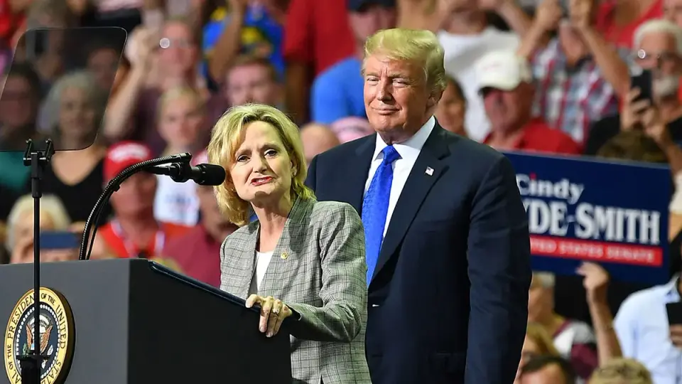 Cindy Hyde-Smith speaks at a campaign podium while Donald Trump stands behind her in an earlier Mississippi rally photograph.