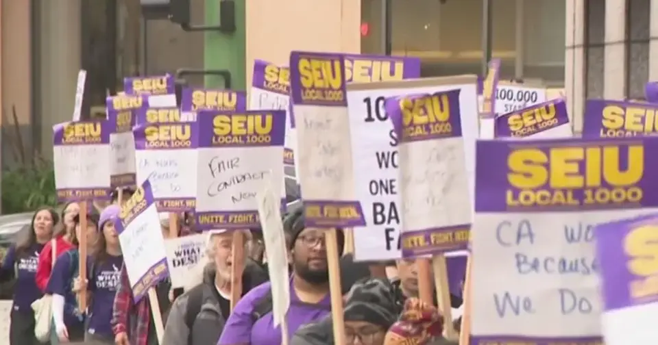 Striking California state workers march on a sidewalk carrying purple and yellow SEIU Local 1000 picket signs during the one-day strike