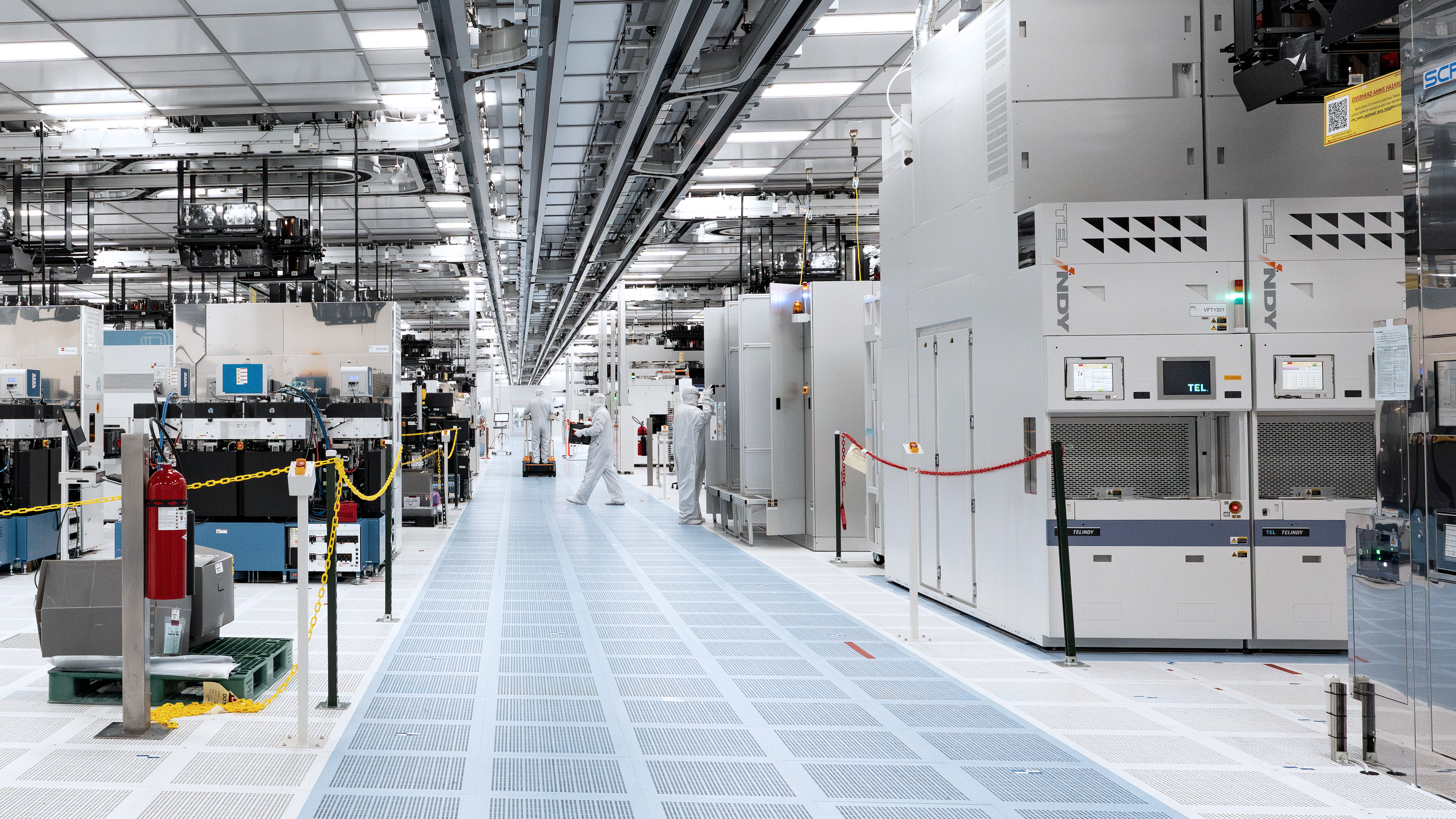 Cleanroom technicians walk between equipment in a semiconductor wafer fabrication facility.