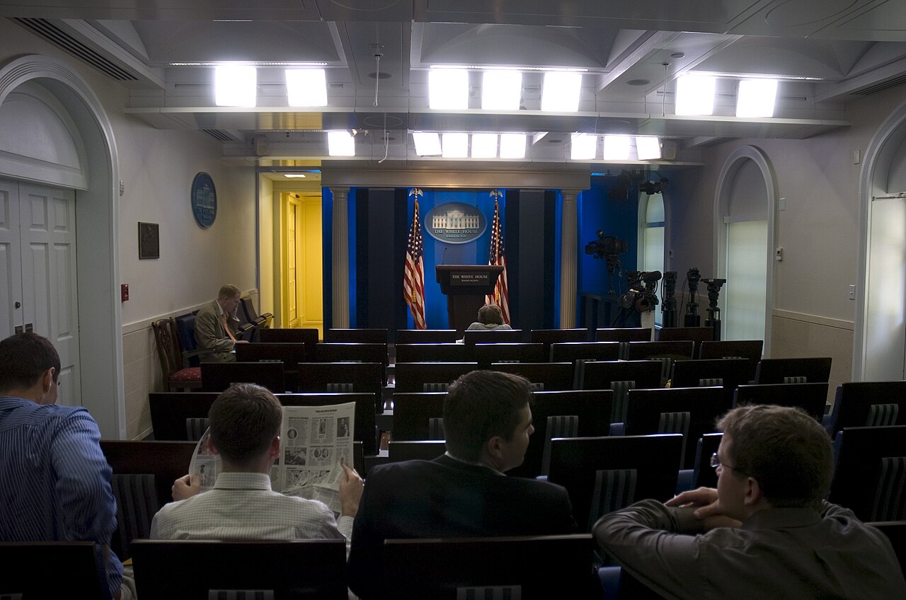 Reporters seated in rows of chairs in the James S. Brady Press Briefing Room at the White House, facing the podium and flags