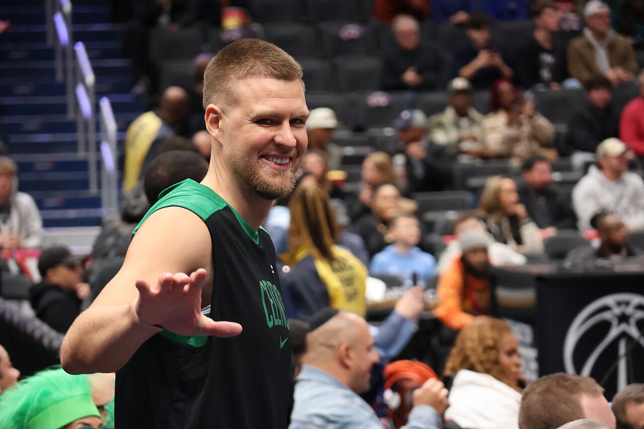 Kristaps Porziņģis smiling and waving toward the camera in a Celtics warm-up shirt before a game in Washington