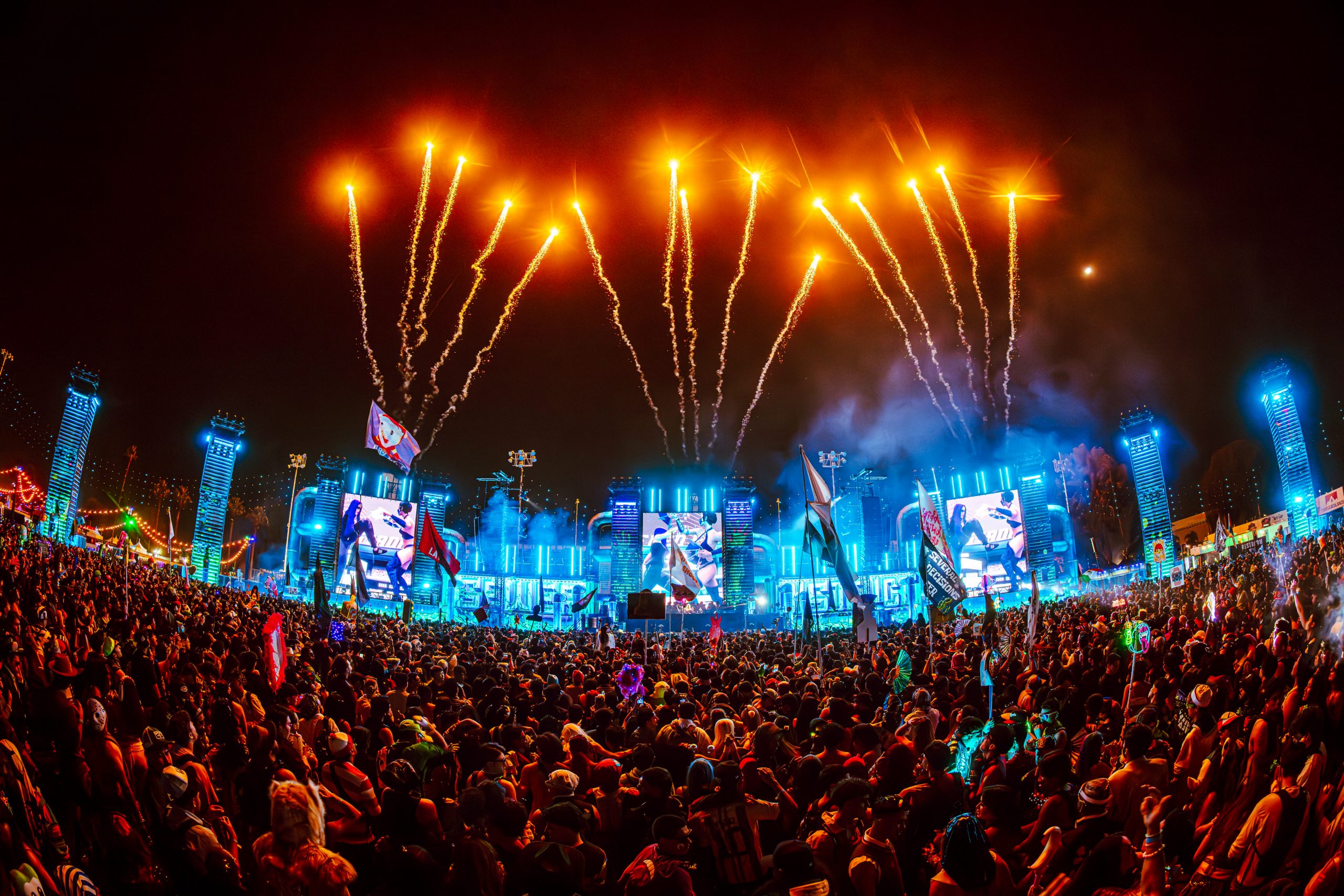 A large crowd in costumes faces an illuminated stage as pyrotechnics fire into the night sky at an Escape Halloween festival
