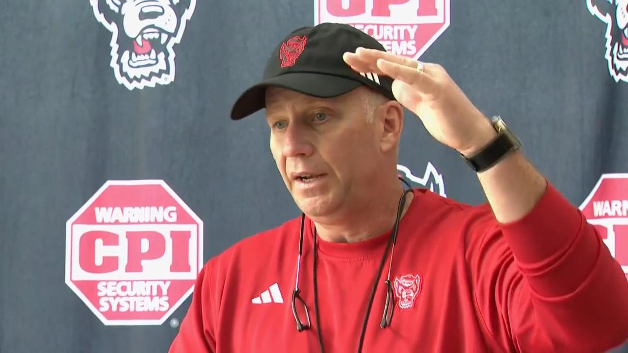 Dave Doeren speaks at an NC State football press conference while wearing a red Wolfpack shirt and black cap