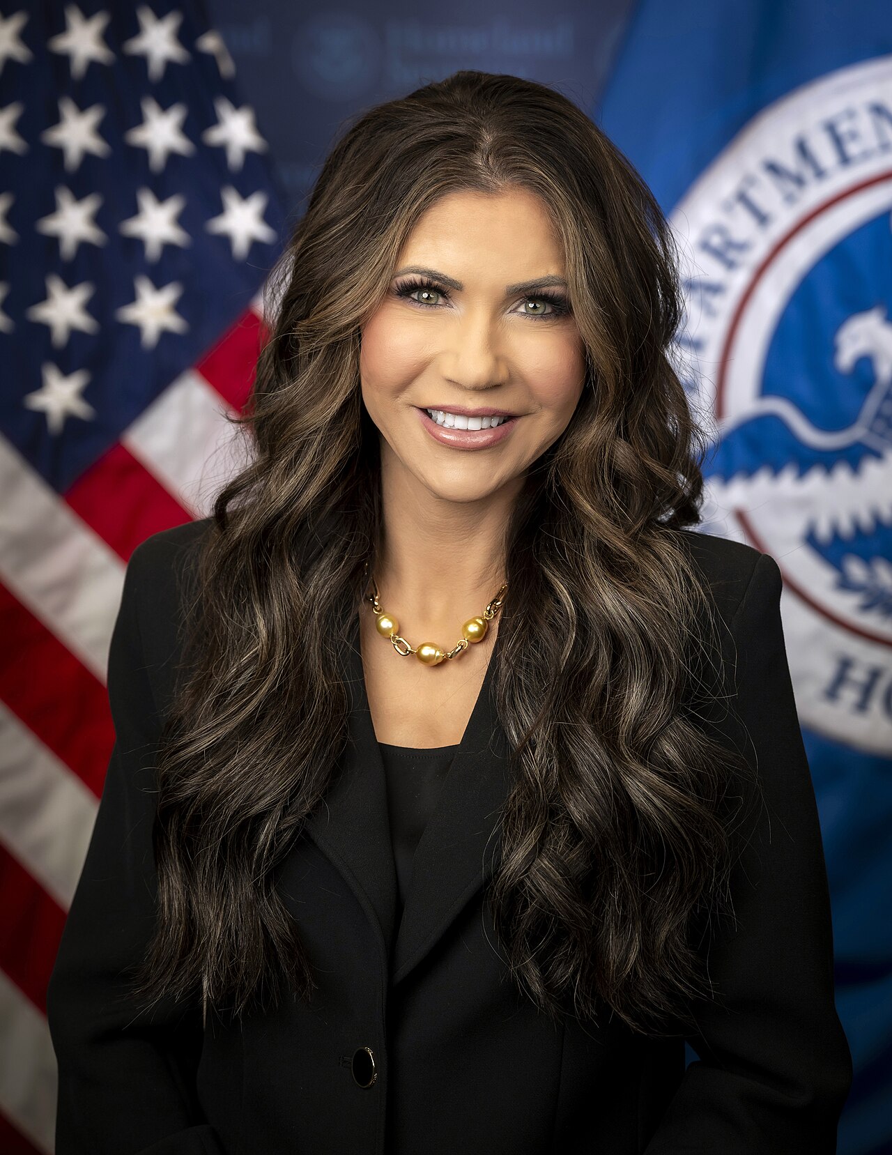 Official portrait of Kristi Noem as U.S. secretary of homeland security, standing in front of American flags